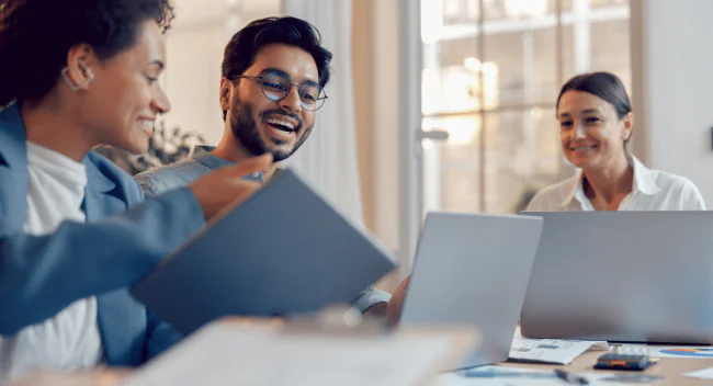 Three colleagues smiling and discussing work around a table with laptops and documents